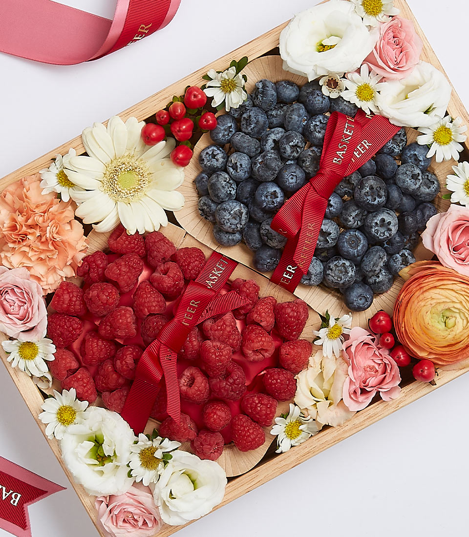 Wooden gift box with fresh raspberries and blueberries, surrounded by white daisies, pink roses, and orange ranunculus