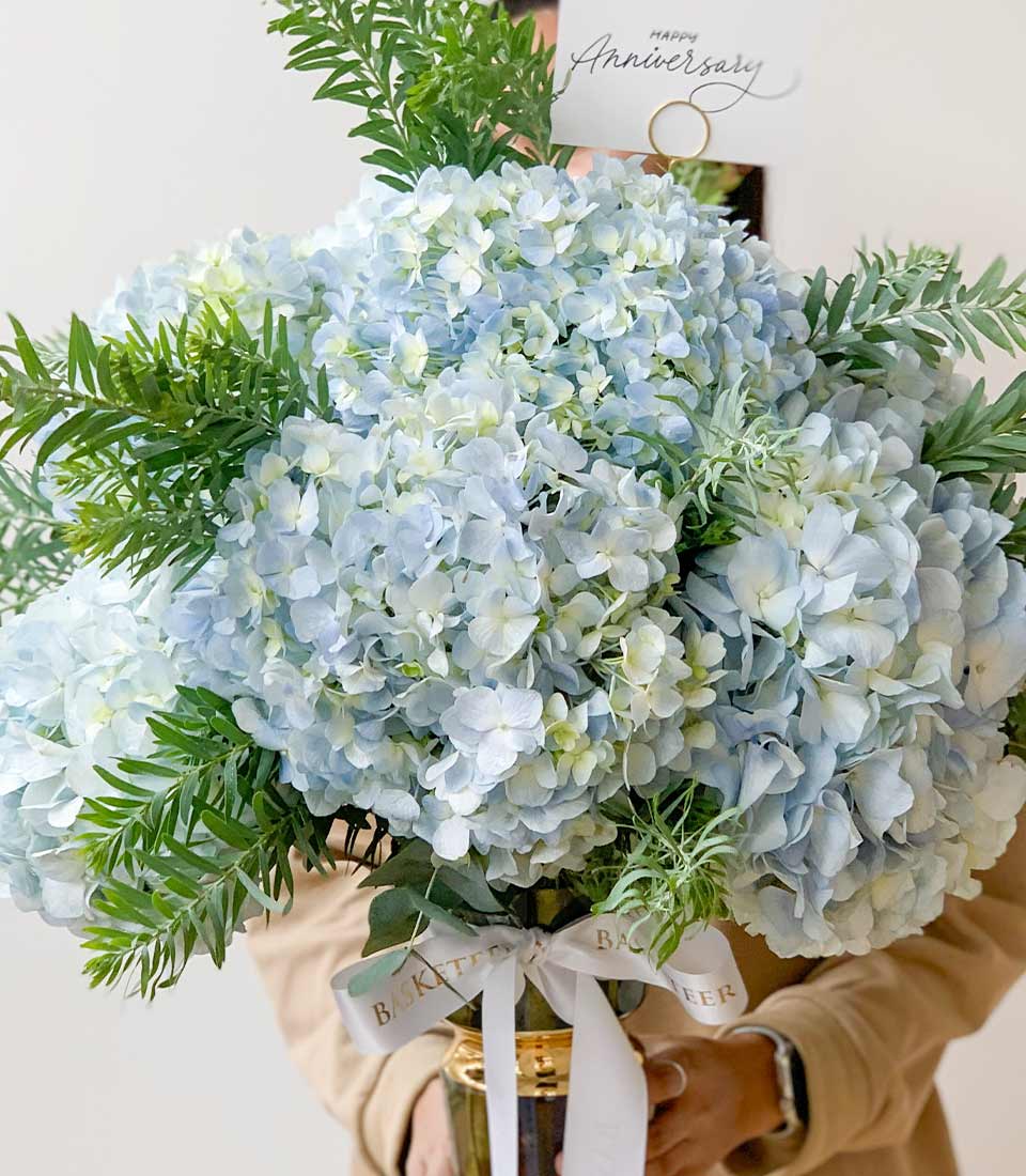 Pastel blue hydrangea bouquet in dark green and gold vase with white ribbon bow