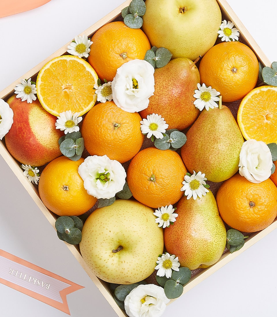 Wooden fruit basket with fresh oranges, apples, pears, white eustoma flowers, and eucalyptus arranged on burlap lining
