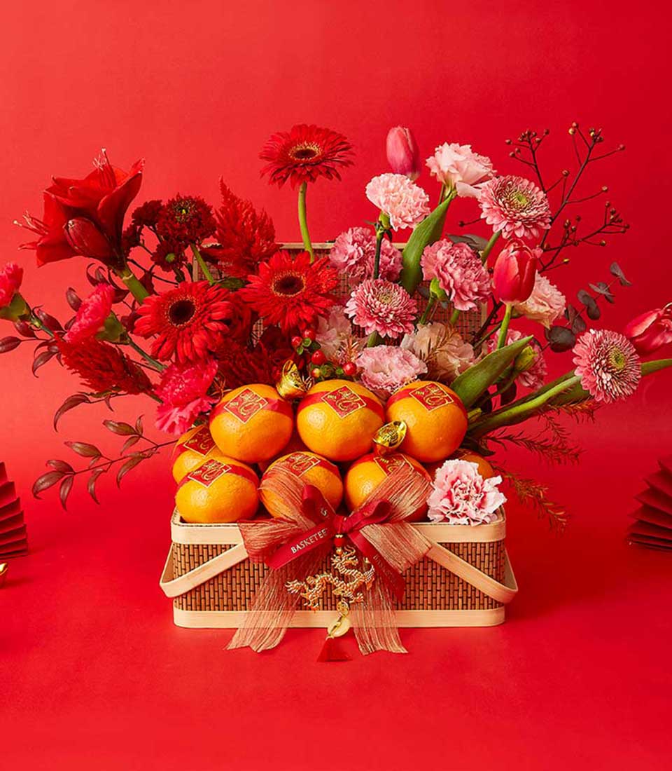 Woven gift basket with fresh red amaryllis, gerbera daisies, pink carnations, and stacked mandarin oranges with gold accents