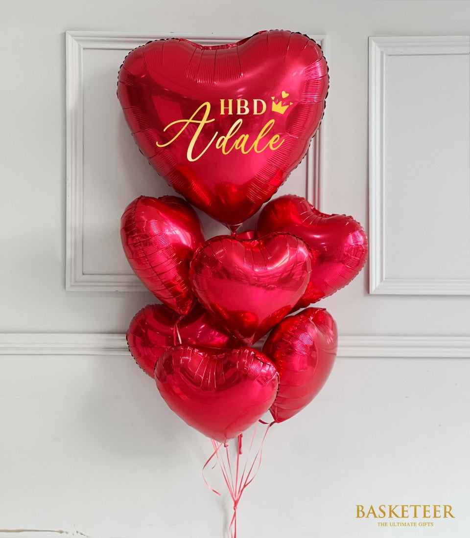Red heart-shaped foil balloons with personalized gold script and crown, tied with red ribbons for Valentine's Day gifts