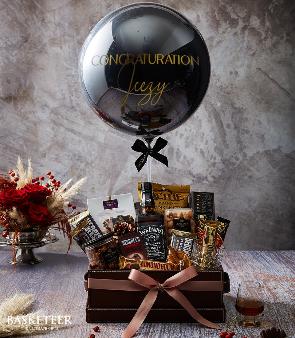 Luxury whisky gift box with chocolates, nuts, gourmet snacks, congratulations balloon, and red rose arrangement