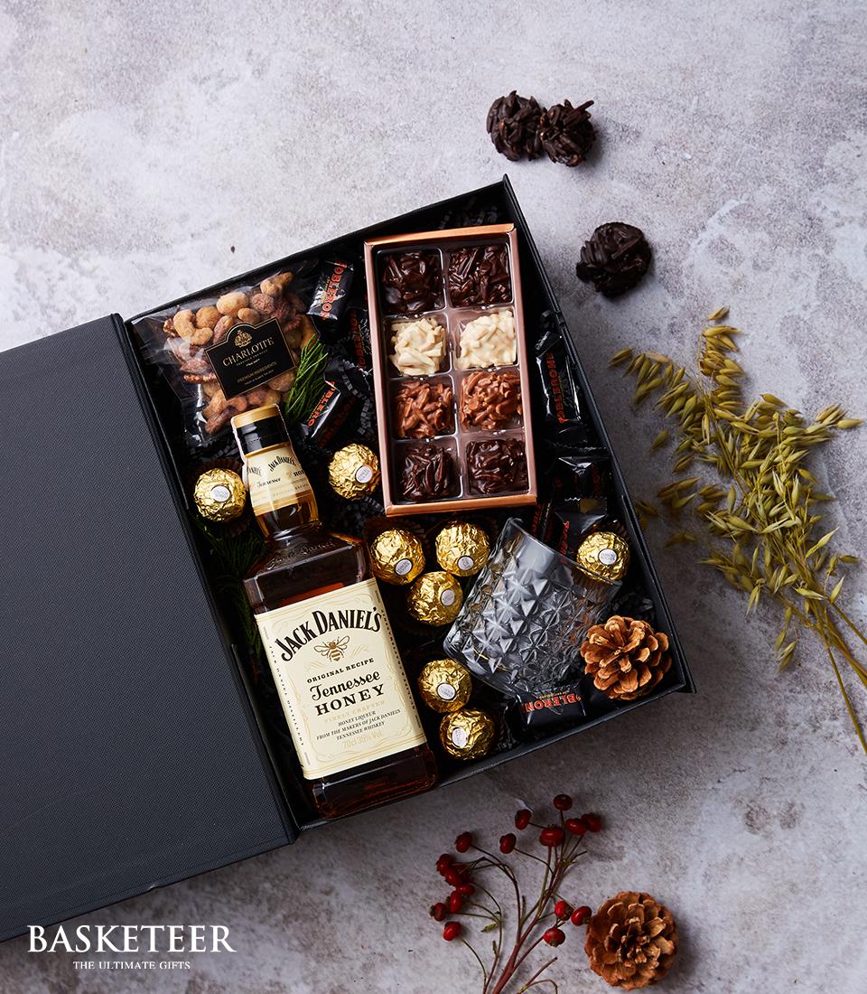 Jack Daniel's Honey whiskey gift box with glassware, nuts, chocolates, and gold-wrapped treats on textured grey surface
