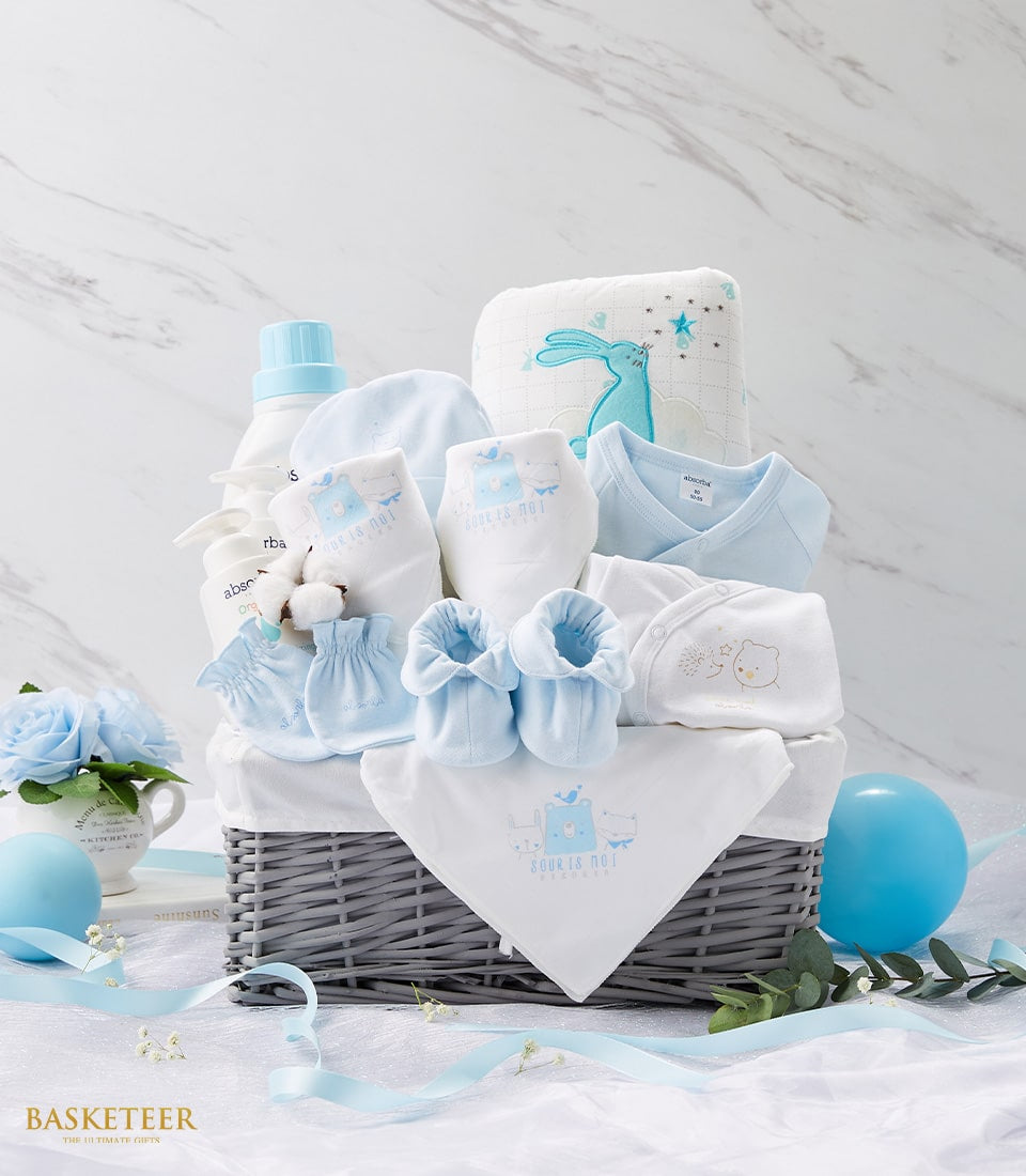 Newborn welcome gift basket with blue onesie, bibs, booties, and baby care products in grey woven basket