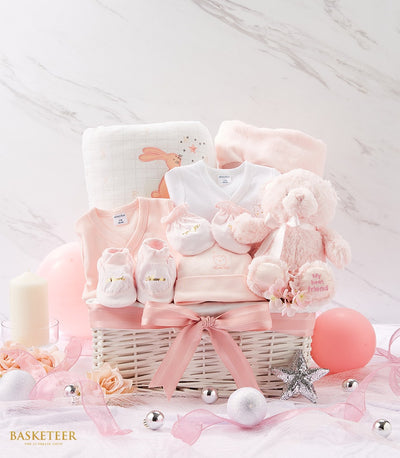 Delicate pink and white newborn gift basket with soft cotton clothing, plush teddy bear, and bunny-patterned blanket