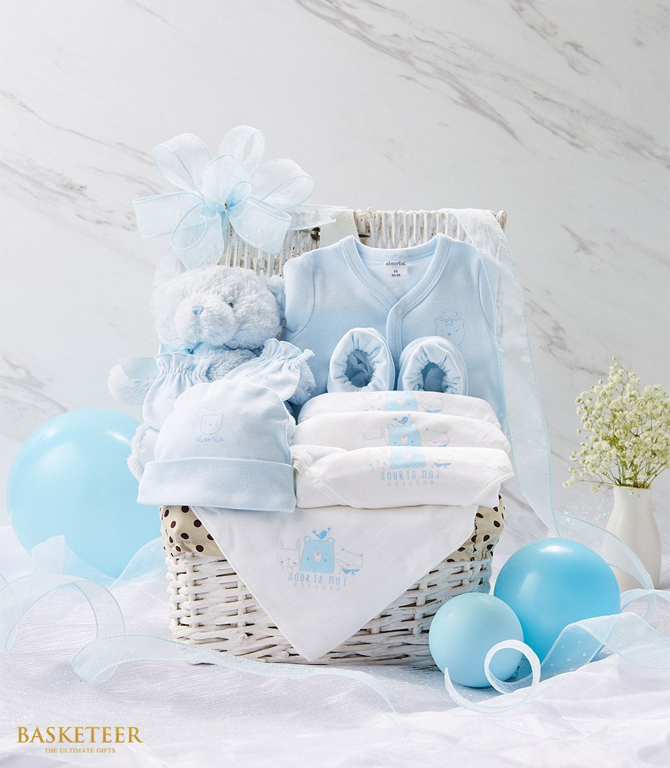 Soft blue newborn gift basket with onesie, hat, booties, teddy bear, and white blankets on woven base