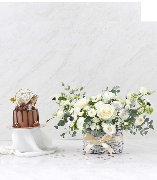 Chocolate layer cake with white flower basket featuring premium roses, ranunculus, and anemones tied with gold ribbon