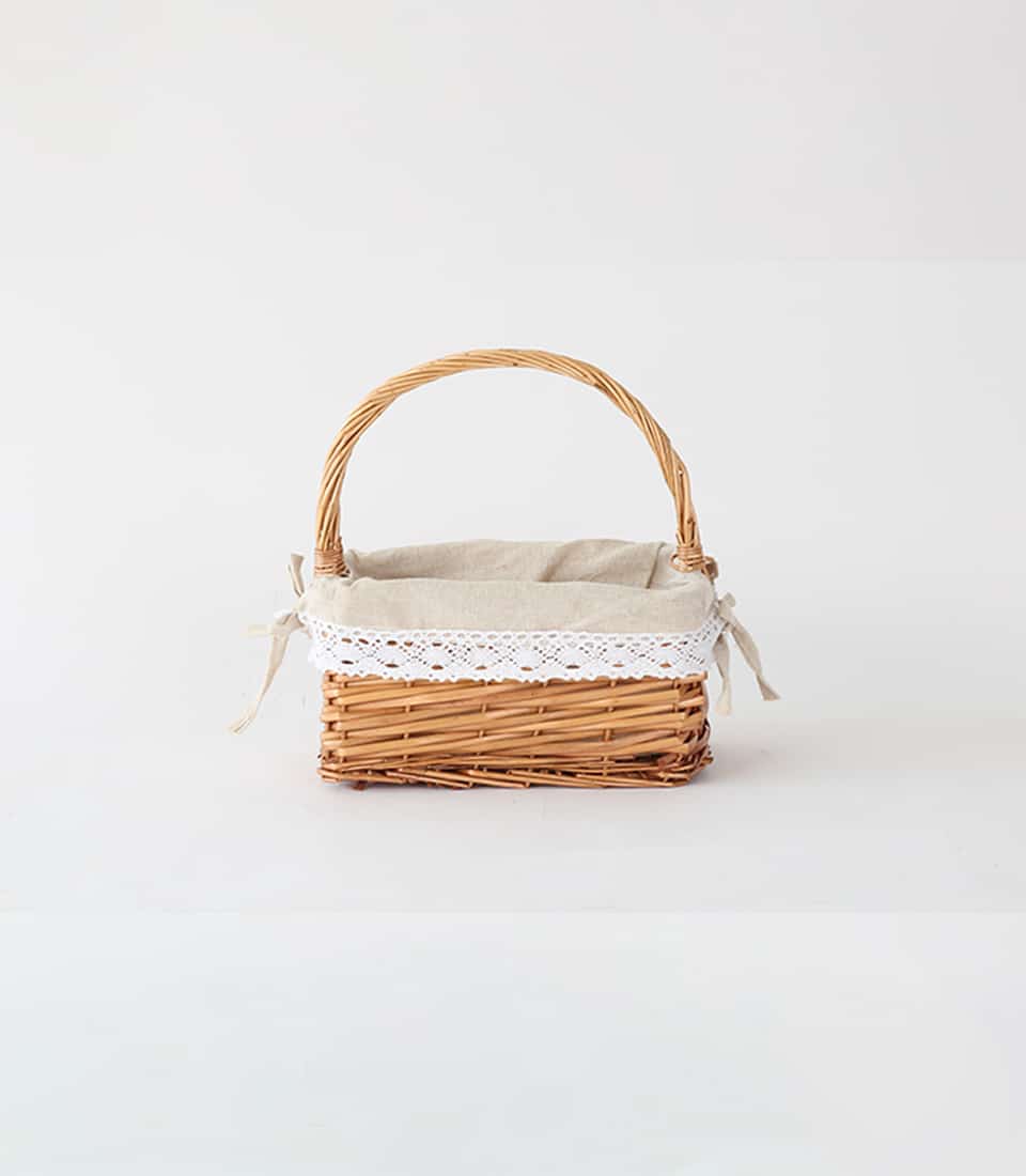 Light brown wicker basket with arched handle, beige fabric liner and white lace trim for gift hamper styling