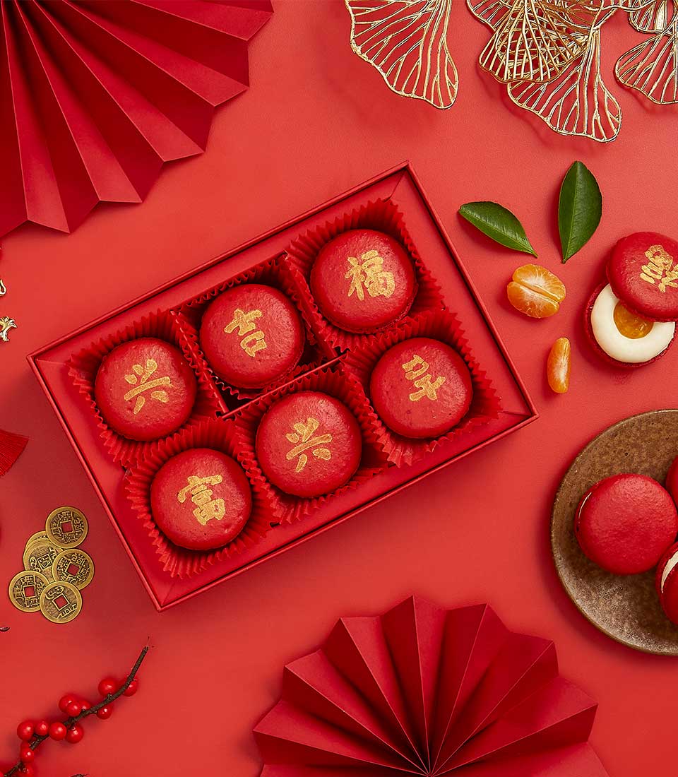 Red macarons with gold Chinese characters in festive CNY gift box with traditional decorations and tangerine accents