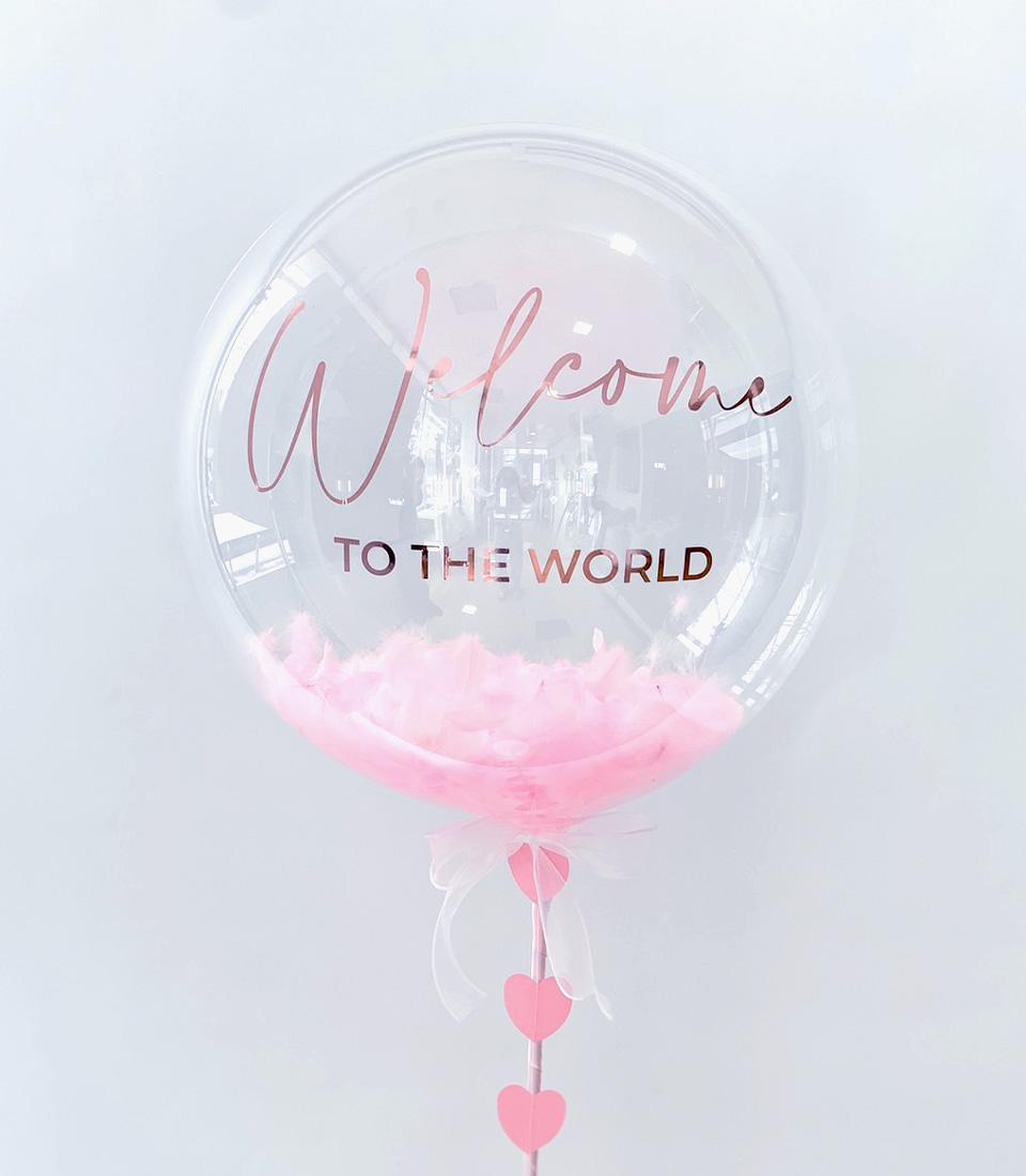 Pink bubble balloon with rose gold welcome message, feathers, and heart charm for new baby gifts