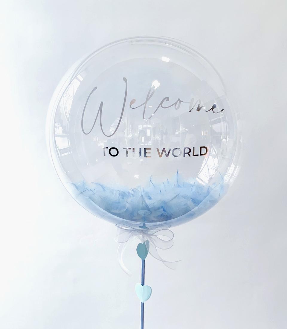 Clear bubble balloon with light blue feathers, silver "Welcome to the World" text, and dangling heart charms on white ribbon