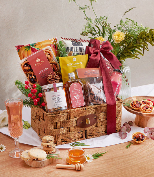 Woven wicker gift basket with gourmet snacks, cookies, honey, almonds, and drinks tied with deep red satin ribbon