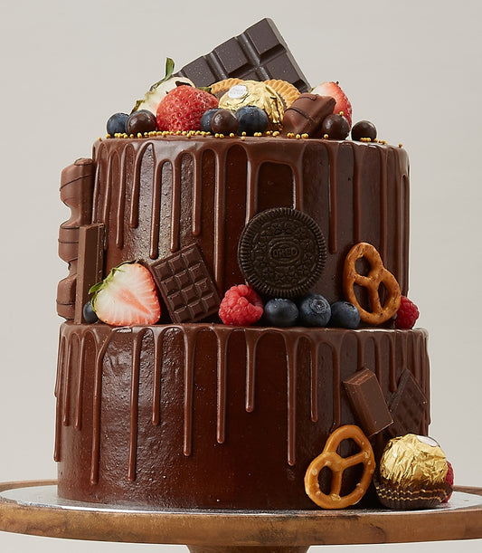 Two-tier dark chocolate drip cake with fresh strawberries, blueberries, chocolate bars, and gold candies on rustic wooden