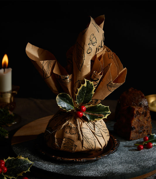 Traditional British Imperial Christmas pudding with holly sprig, brown paper wrapping, and dark fruit slice on ornate plate