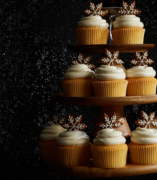 Twelve vanilla cupcakes with white frosting and gingerbread snowflake cookies on tiered wooden stand, festive Christmas gift