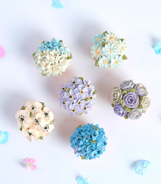 Six delicate floral cupcakes with buttercream hydrangeas, roses, and daisies in pastel blues and purples for Mother's Day
