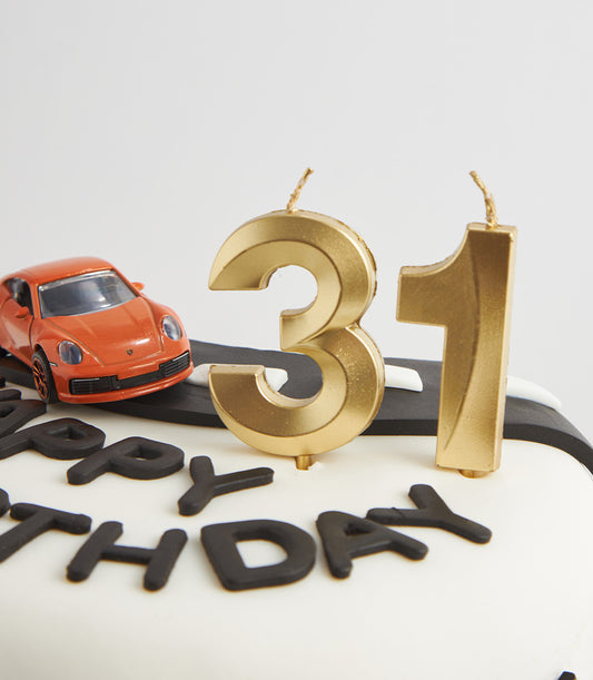 White fondant birthday cake with black road design, toy sports car, and golden numeral candles for 31st celebration