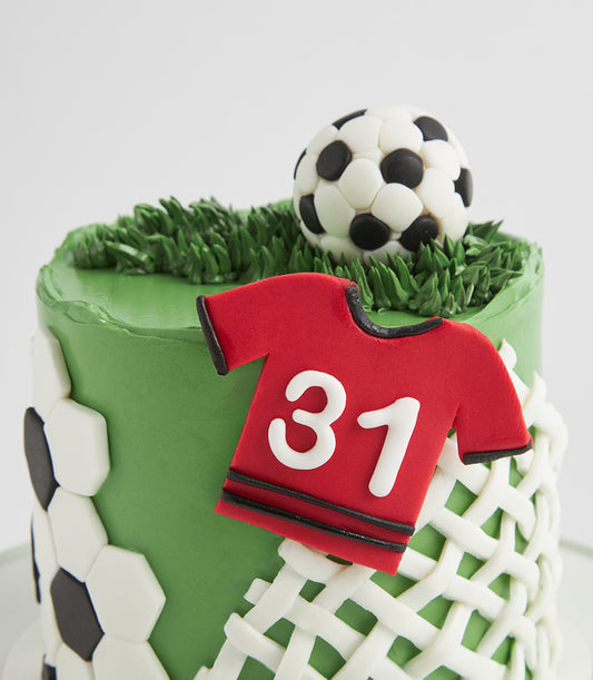 Two-tier soccer birthday cake with grass-textured green frosting, edible ball, net pattern, and red jersey with number 31