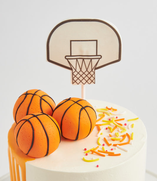 White frosted birthday cake with basketball hoop topper and orange candy basketballs, colorful sprinkles