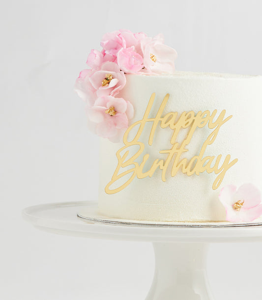 White layer cake with cascading pink flowers and gold Happy Birthday topper on silver board