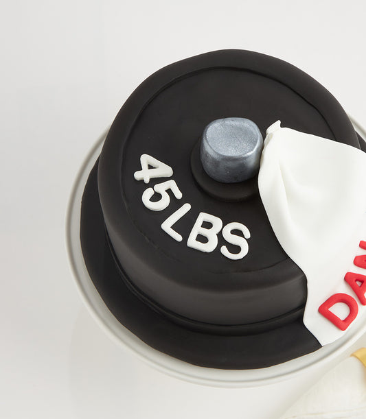 Black fondant barbell weight plate cake with white "45 LBS" lettering and silver collar, designed for fitness enthusiast
