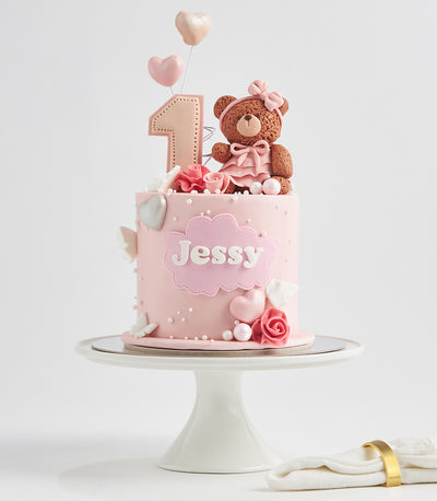 Light pink first birthday cake with teddy bear, hearts, and personalized name plaque on ceramic stand