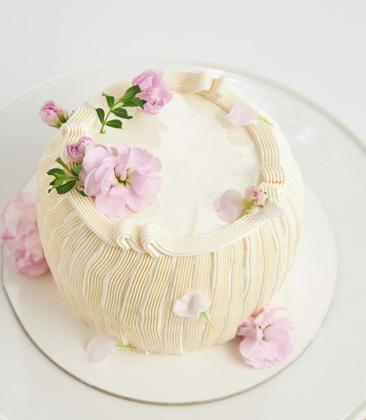 Lemon cream layer cake with ridged frosting, fresh pink flowers, and delicate piping on white stand