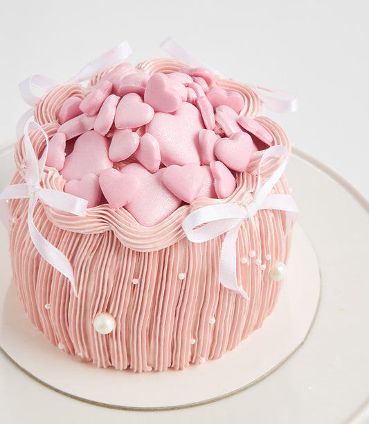 Soft pink layer cake with ruffled frosting, heart-shaped candies, white ribbon bows, and pearl decorations for birthdays