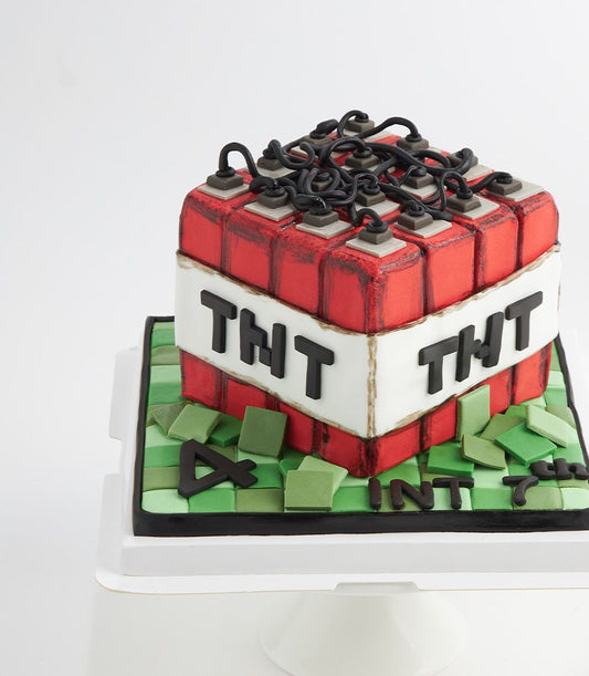 Square red Minecraft TNT birthday cake with white band, black wires, grey detonators, and grass block base for kids'
