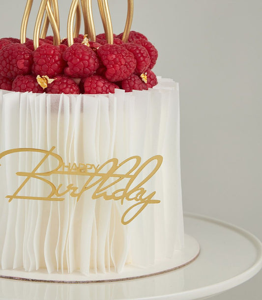 Elegant white layer cake with fresh raspberries, gold leaf, and script birthday topper