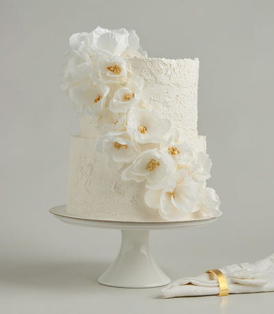 Two-tier white wedding cake with cascading wafer paper flowers and gold leaf detail on rustic buttercream frosting