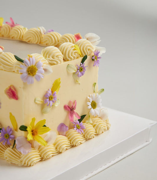 Personalised square layer cake with pale yellow buttercream, edible flowers, and delicate piped borders