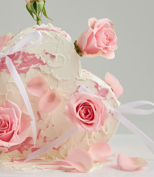 Heart-shaped strawberry cake with pink roses, white textured frosting, and rose petals for romantic gifting