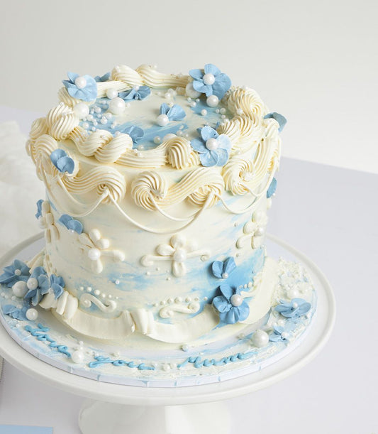 Two-tier Mother's Day cake with white buttercream, light blue watercolor swirls, and pearl-adorned sugar flowers