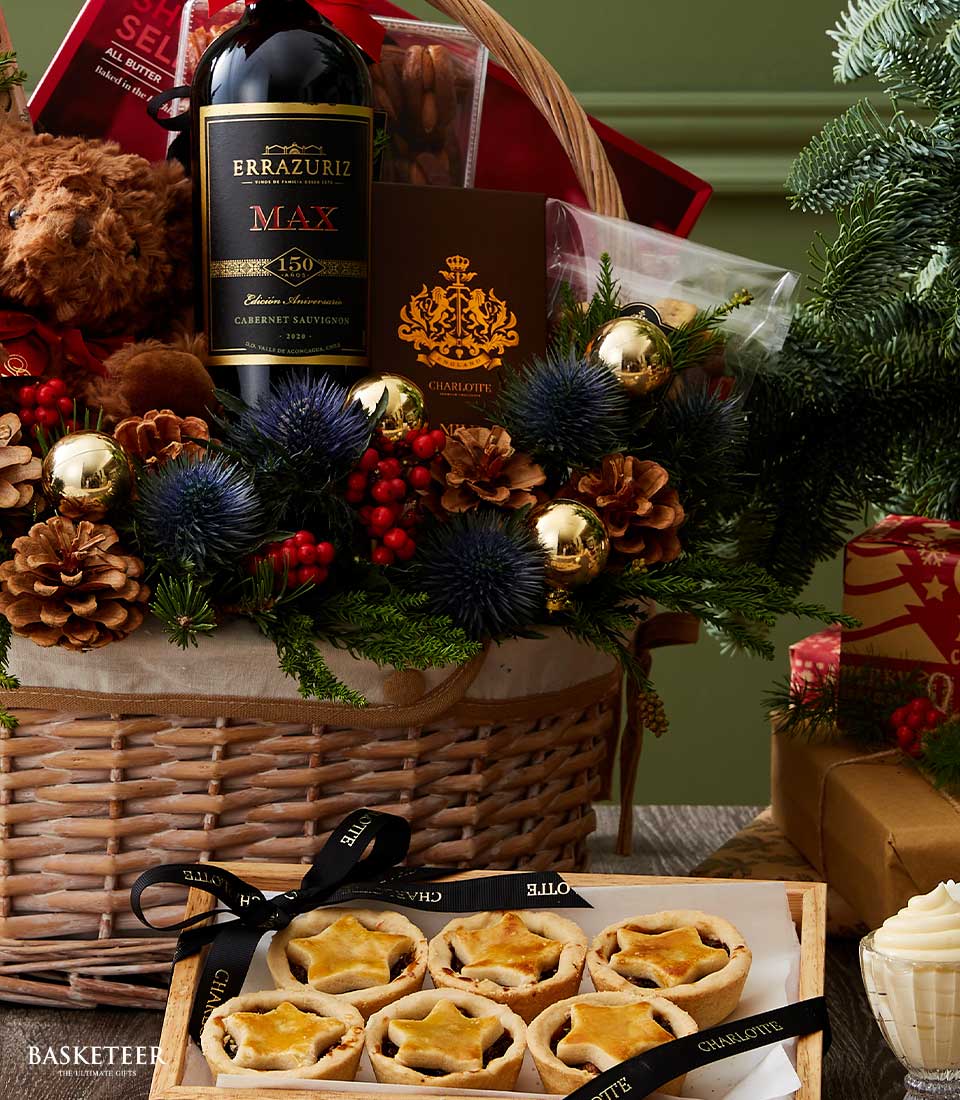 Festive wicker hamper with red wine, mince pies, teddy bear, and holiday florals for Christmas gifting