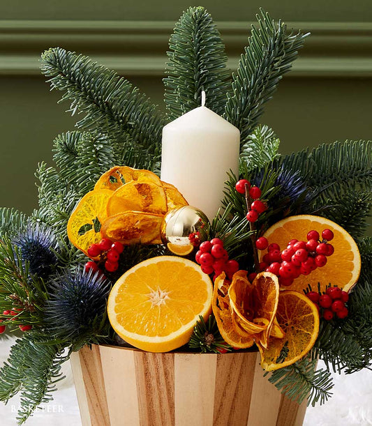 Rustic citrus Christmas centerpiece with white pillar candle, fresh fir, orange slices, and red berries in striped wood
