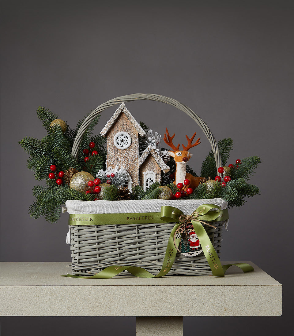 Festive grey wicker Christmas basket with reindeer figurine, gingerbread houses, gold baubles, and pine branches