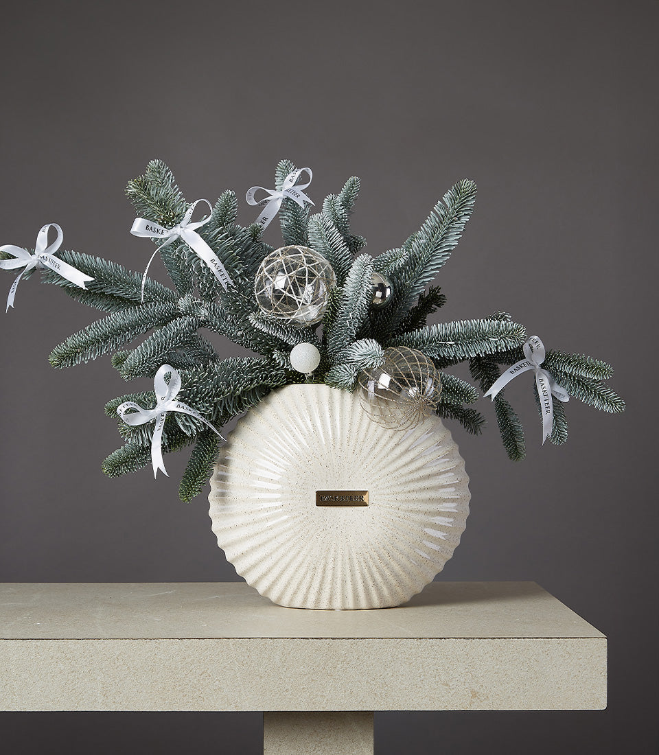 Fresh evergreen and frosted pine arrangement in cream ceramic vase with glass baubles and white ribbon bows, festive