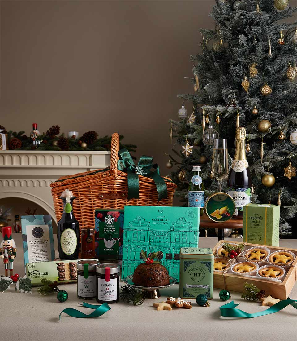 Green ribbon wicker Christmas hamper with sparkling wine, champagne flutes, Christmas pudding, and gourmet holiday treats