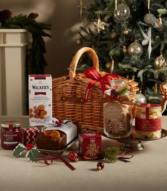 Woven wicker Christmas hamper with shortbread, gingerbread, jam, fruit cake, and festive tea, tied with red ribbon