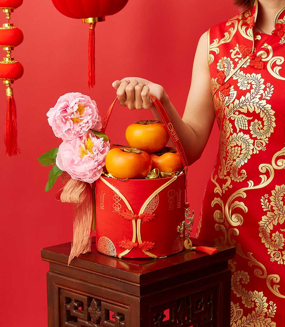 Lunar New Year gift box with persimmons and gold ingots, pink peonies, traditional red cheongsam styling on carved wooden