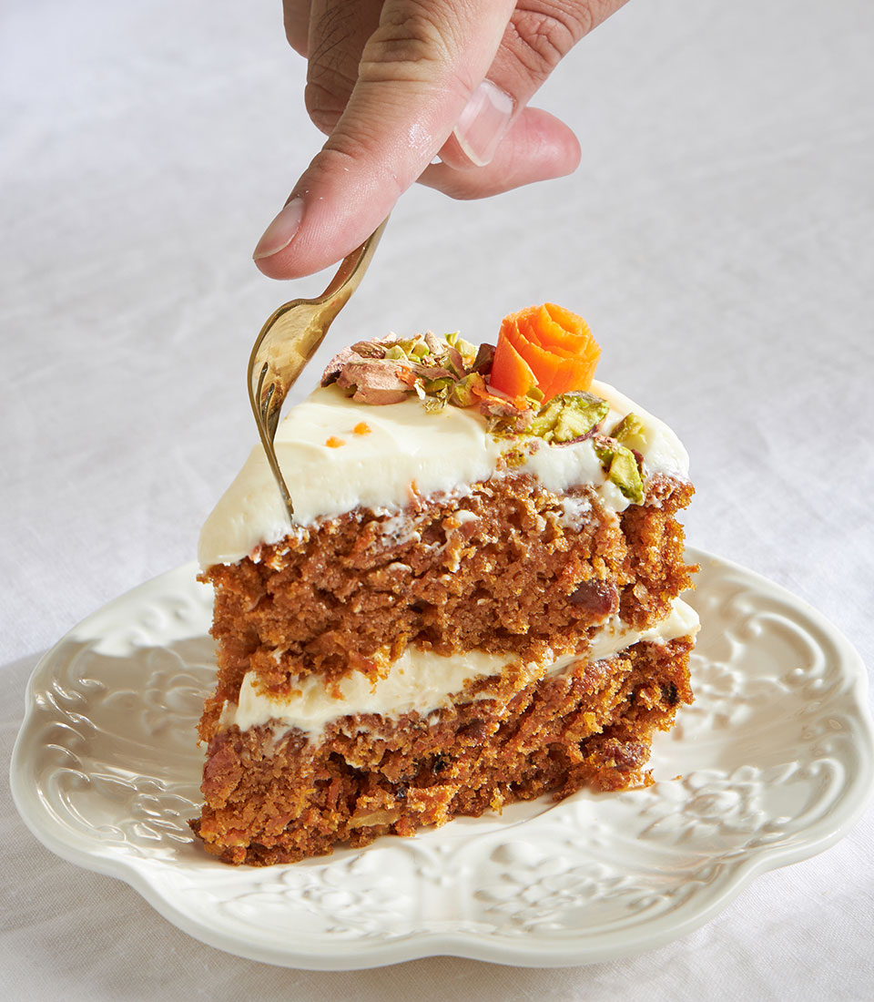 Layered carrot cake with cream cheese frosting, pistachio, and candied carrot garnish on scalloped plate