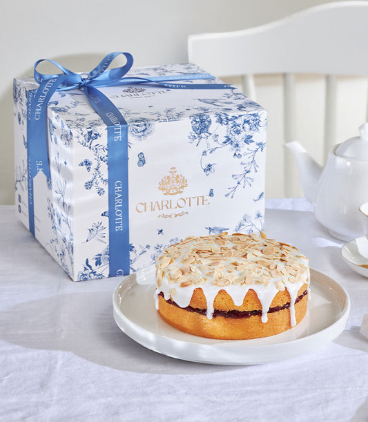 Cherry Bakewell almond cake with white icing drip and sliced almonds, presented in blue floral gift box with satin ribbon