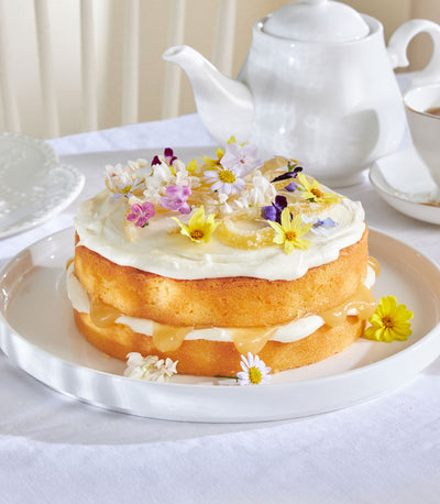Two-tier lemon drizzle cake with white frosting, caramel glaze, edible flowers and candied lemon on ceramic plate