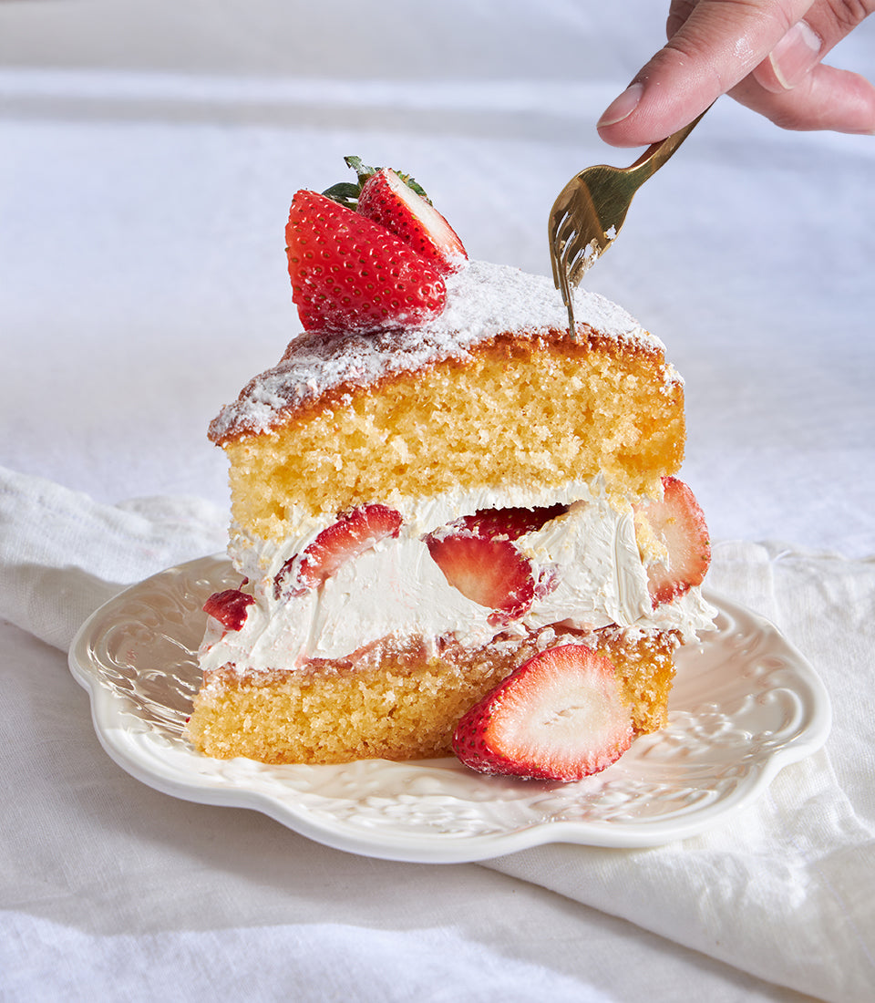 Thai-style strawberry Victoria cake slice with golden sponge, fresh cream, and powdered sugar on a scalloped plate