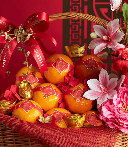 Wicker basket overflowing with mandarin oranges tied with red ribbons, golden ingots, and pink-white artificial flowers for