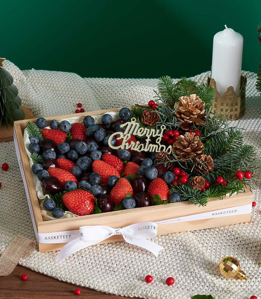 Wooden gift box with fresh strawberries, blueberries, and cherries, festive pine branches, and gold "Merry Christmas"
