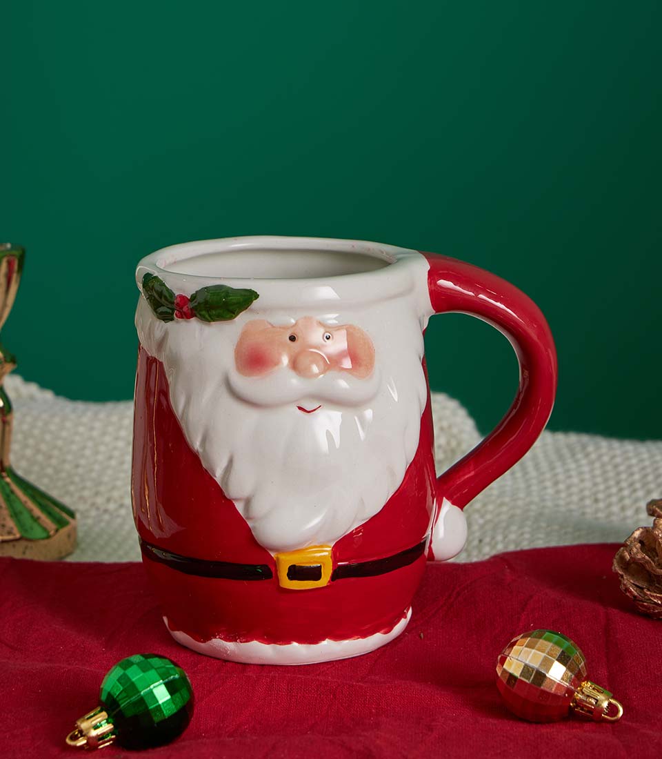Ceramic Santa mug with red and white design, green holly, and gold buckle on festive red fabric with Christmas ornaments
