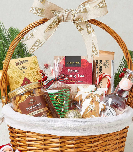 Woven Christmas hamper with chocolate, tea, popcorn, and festive treats in cream wicker basket with gold ribbon