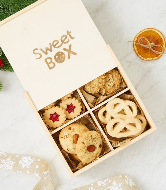 Wooden gift box with assorted gourmet cookies, jam-filled stars, almond rounds, and butter pretzels on marble with holiday